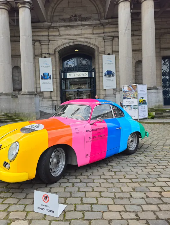 A vintage Porsche painted in rainbow stripes, parked on cobblestones outside Autoworld