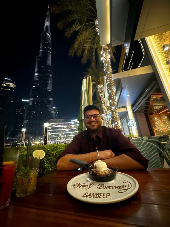 Sandeep at a restaurant table with a birthday dessert, the Burj Khalifa lit up behind him