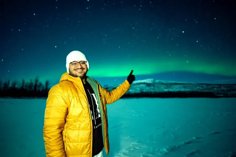 Sandeep in a yellow puffer jacket on a snowfield at night, pointing at green northern lights