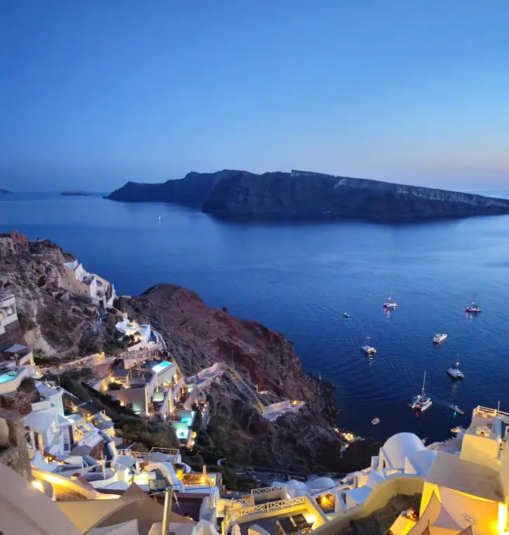 Oia's white houses on the cliff at blue hour, with boats on the sea below