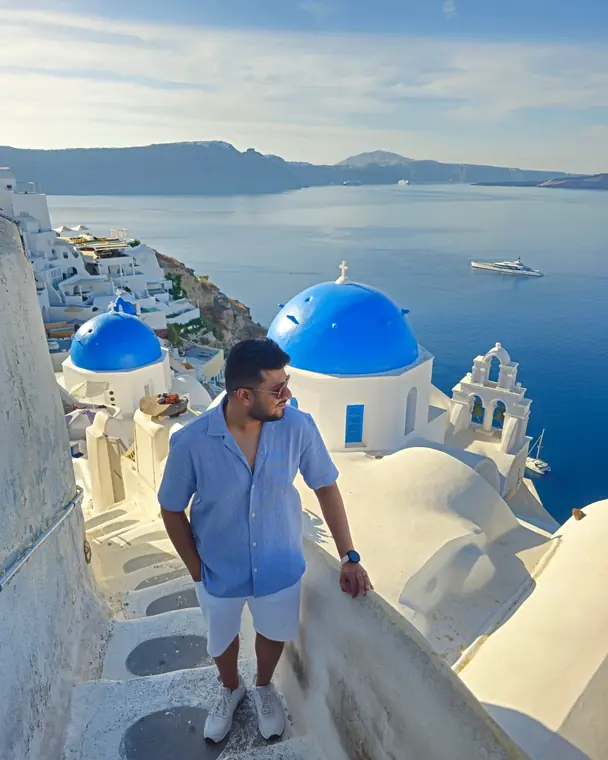 Sandeep on a whitewashed terrace in Oia beside blue-domed churches, with the sea behind
