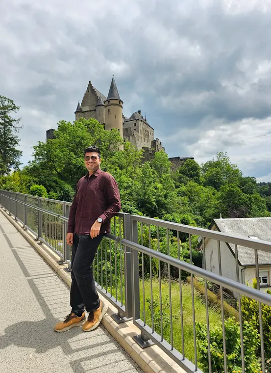 Sandeep on a footpath, with Vianden Castle rising above the trees behind him